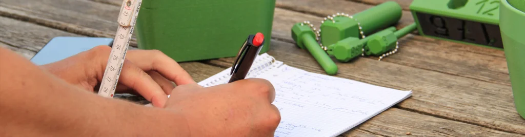 Detailaufnahme einer Spielplatzprüfung: Hände mit Kugelschreiber schreiben etwas auf einen Block. Im Hintergrund Prüfkörper gemäß DIN EN 1176