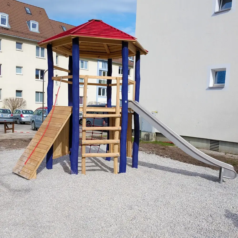 Sechseckiger Spielturm mit blauen Standpfosten, rotem Spitzdach, einer Rampe mit rotem Seil, einer Sprossenleiter und einer Edelstahlrutsche. Der Turm steht auf einer Kiesfläche zwischen mehrstöckigen Wohnblocks