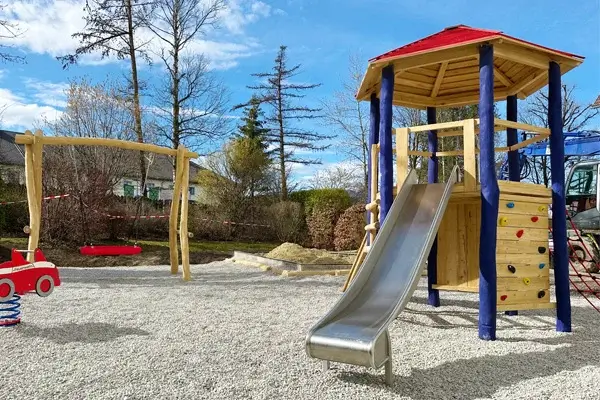 6Eck-Turm aus Robinie mit blauen Standpfosten mit einer Kletterwand mit bunten Klettergriffen, einer Edelstahlrutsche und weiteren Auf- und Abstiegen. Das Gerät steht auf einer Fläche aus Fallschutzkies. links daneben ein einfaches Federwippfahrzeug in rot und im Hintergrund eine rote Nestschaukel