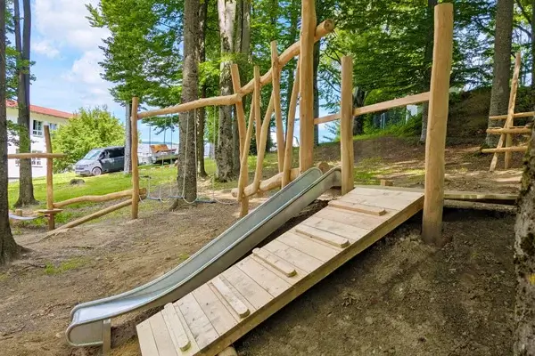 Motorikpfad im Wald: In einem Buchenwaldstück im Vordergrund eine Aufstiegsrampe aus Robinienholz an einem Hang. direkt daneben eine Edelstahl-Hangrutsche. Im Hintergrund verläuft ein Motorikpfad aus Robinienstämmen mit verschiedenen Spielelementen