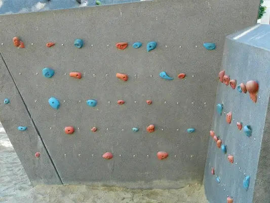 kleine, niedrige Boulderwand auf Spielplatz. Graues Eck aus Beton mit roten und blauen Klettergriffen. 