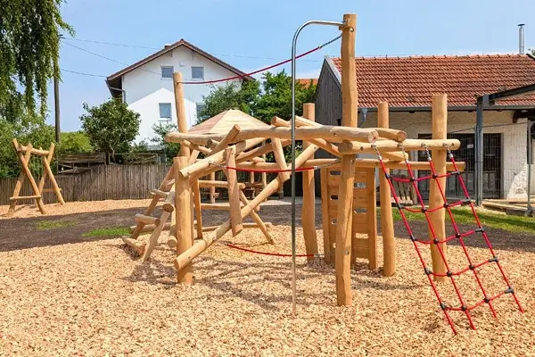 KiTa Außengelände: Im Vordergrund ein Klettergerüst aus Robinienstämmen mit rotem Kletternetz und einigen Handlaufseilen. Im Hintergrund links am Bildrand: eine Doppelschaukel mit Robiniengestell. Dahinter ein Bretterzaun und ein Geräteschuppen.