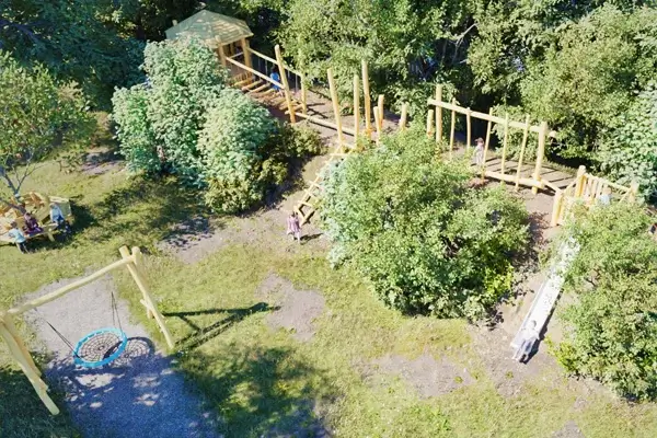 schräge Ansicht von oben auf einen langgestreckten, künstlichen Hügel im AUßenbereich eines Kindergartens. Der Hügel ist an den Flanken mit Bäumen und Büschen bewachsen. Auf dem Hügel erstreckt sich ein Spiel-Motorikpfad aus Robinienholz. An beiden Enden ist ein Spielturm zu erkennen, der hintere mit Dach. Nach vorne läuft eine schräge Leiter am Hügel nach oben zum Motorikpfad, mit etwas Abstand führt eine Rutsche in die selbe Richtung von oben herunter.