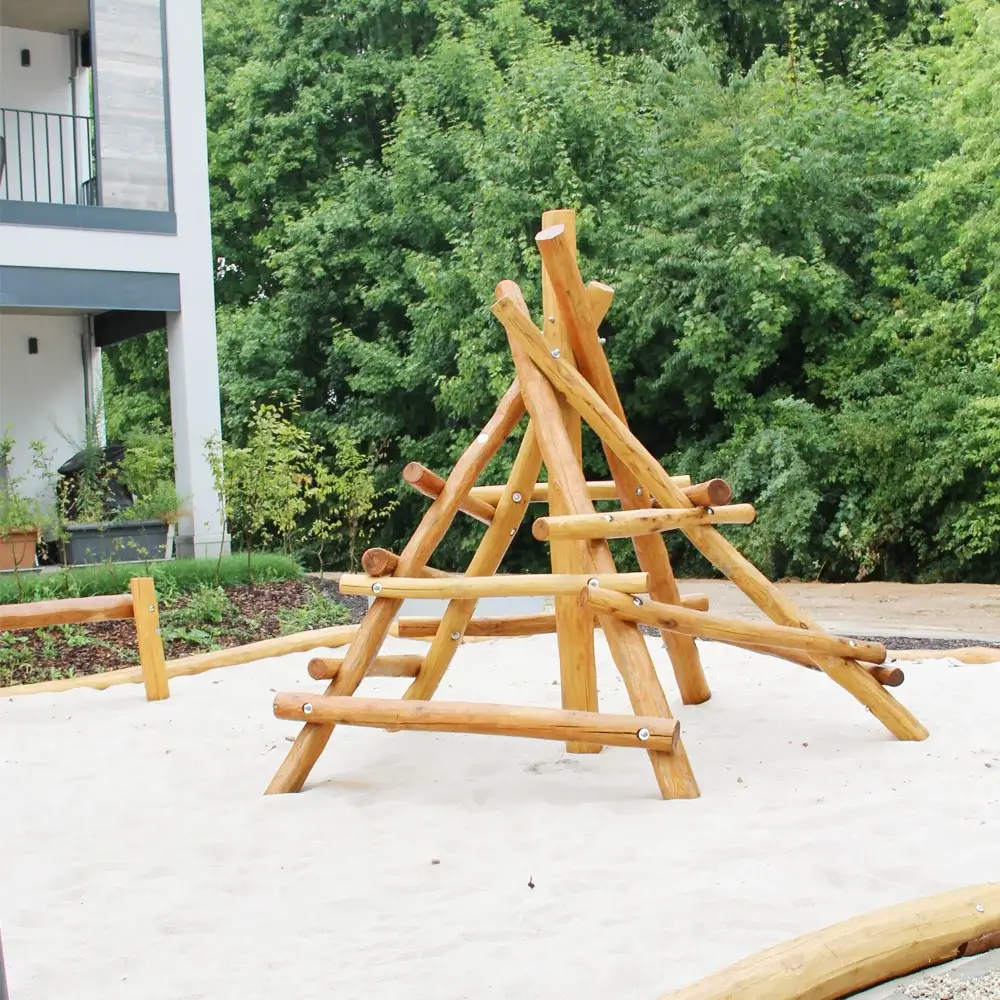 Klettergerüst Pyramide auf Fallschutzsand-Fläche. Im Hintergrund eine grüne Laubhecke und angeschnitten die Ecke eines Wohnhauses. Teil von Wohnquartier Spielplätze