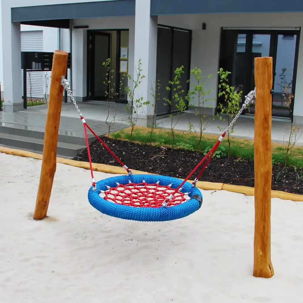 Eine Mini-Nestschaukel mit rotem Netz und blauer Umrandung, niedrig aufgehängt an zwei Robinienpfosten, steht auf weißem Sand. Im Hintergrund eine leichte, begrünte Böschung zu einer Terrasse einer Wohnung