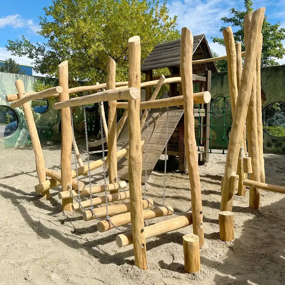 individueller Motorikpfad: Niedrigseilgarten als Kreisförmige Erweiterung eines Bestand-Spielturms auf einer Sandspielfläche. Der Turm ist als zentrales Element Start- und Endpunkt des Parcours zugleich. Im Vordergrund, im Bild, eine Wackelbrücke aus hängenden Trapezförmigen Robinienknüppeln