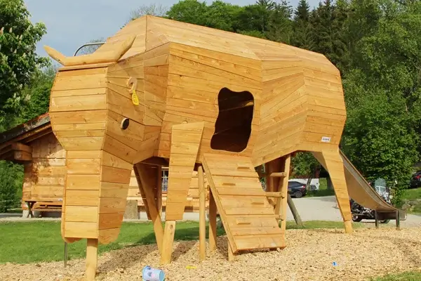 Spielanlage Holzkuh von vorne gesehen. Der Kopf der Kuh neigt sich zum Boden zum Fressen. Seitlich im Bauch ist eine Öffnung in Form eines Kuhfleckens durch den Kinder über eine Rampe ins Innere klettern können. Am Hintern führt eine Rutsche wieder ins Freie