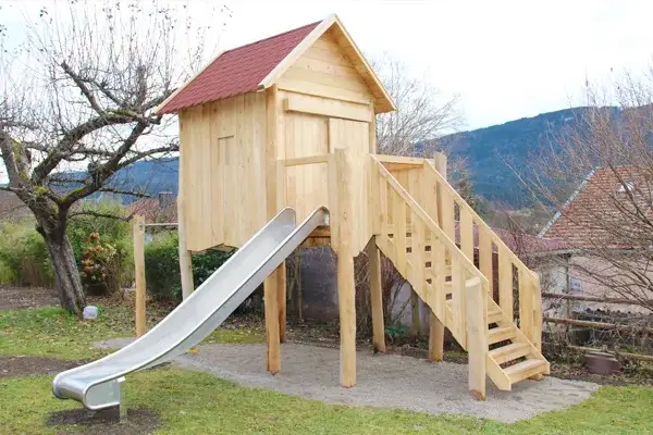 Stelzenhaus aus Robinie in einem privaten Garten mit kleinem Balkon mit Zugangstreppe, sowie einer Edelstahlrutsche. Das Dach ist mit roten Bitumenschindeln gedeckt.