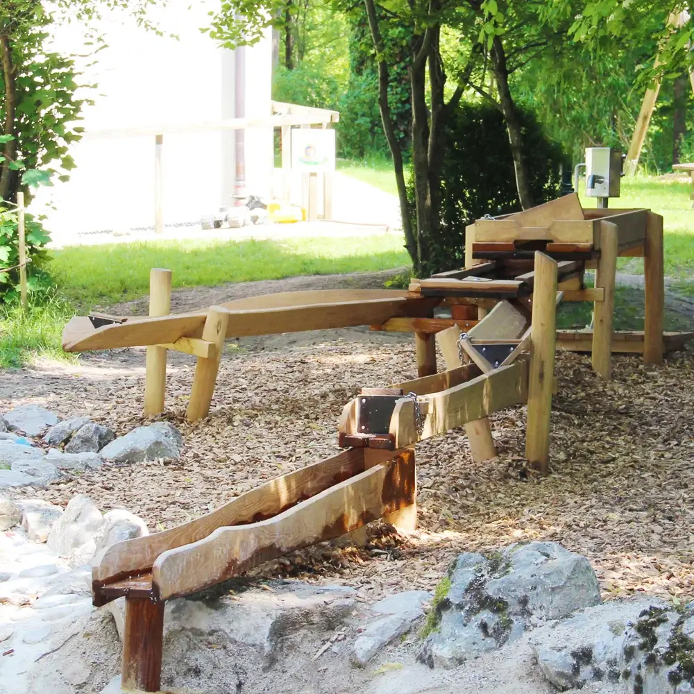 individueller Wasserlauf aus Robinienrinnen und -staukästen bestehend aus mehreren, hintereinander gestaffelten Wasserrinnen, teilweise mit Staubrettern. Mittig eine Gabelung der Rinnen. Im Hintergrund, hinter einem größeren Staubecken ist eine Edelstahlpumpe erkennbar.