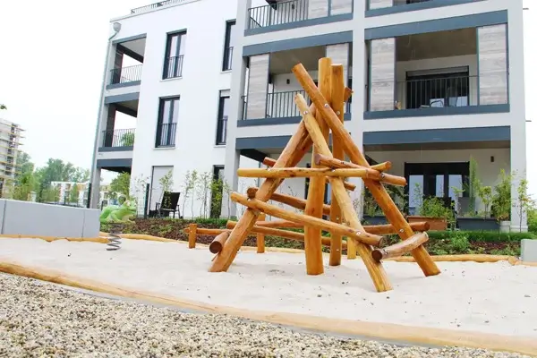 Ansicht eines Abschnitts von mehrern Wohnquartier Spielplätzen. Foto von Bodennähe nach schräg oben. Im Vordergrund ein Klettergerüst aus Robinienstämmen, pyramidenförmig. Dahinter auf der selben Fallschutz-Sandfläche ein grüner Federwippfrosch. Im Hintergrund ein mehrstöckiger, moderner Neubau